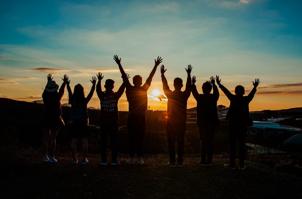 7 junge Menschen die mit dem Rücken zur Kamera in einen Sonnenuntergang schauen und dabei die Hände in die Luft strecken