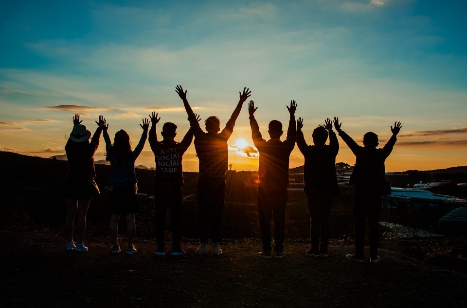 7 junge Menschen die mit dem Rücken zur Kamera in einen Sonnenuntergang schauen und dabei die Hände in die Luft strecken