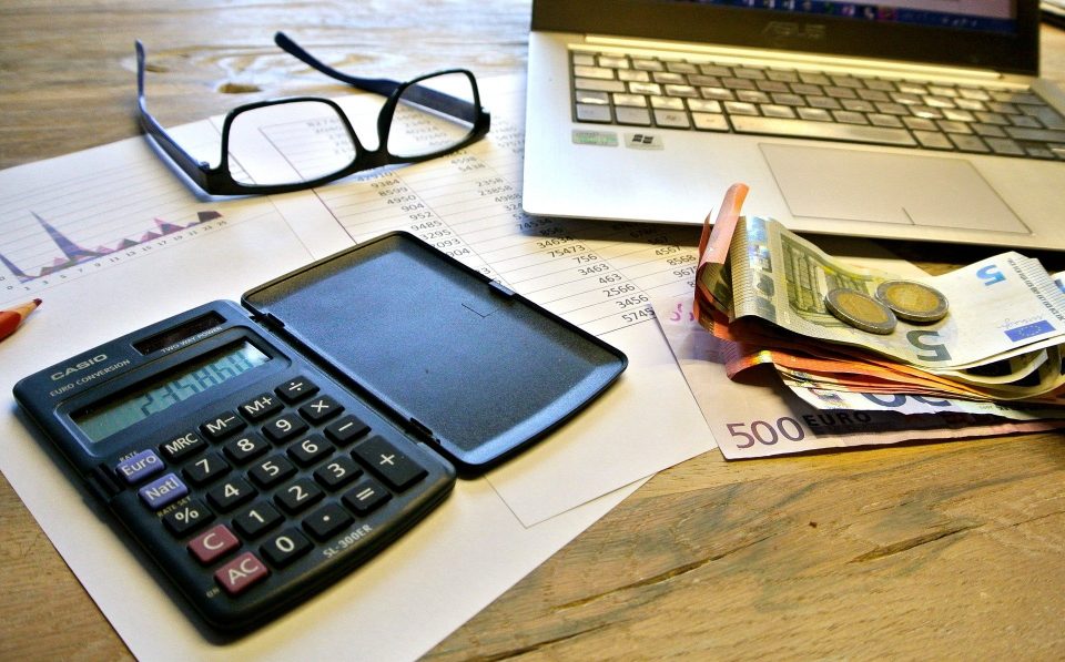 Auf einem Tisch liegen Taschenrechner, eine Brille, ein Laptop und Geld.