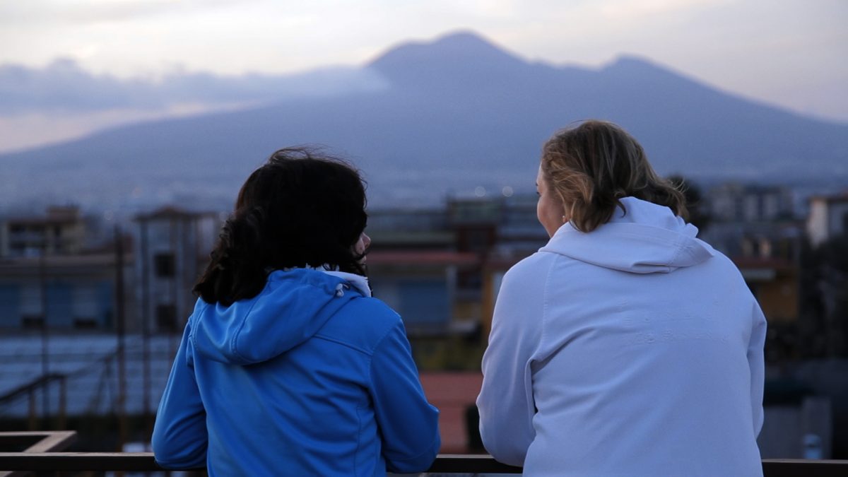 Das Bild zeigt eine Filmszene, auf der zwei Frauen von hinten zu sehen sind, die im Gespräch zu sein scheinen. Im Hintergrund ist eine Landschaft mit Berg zu sehen.