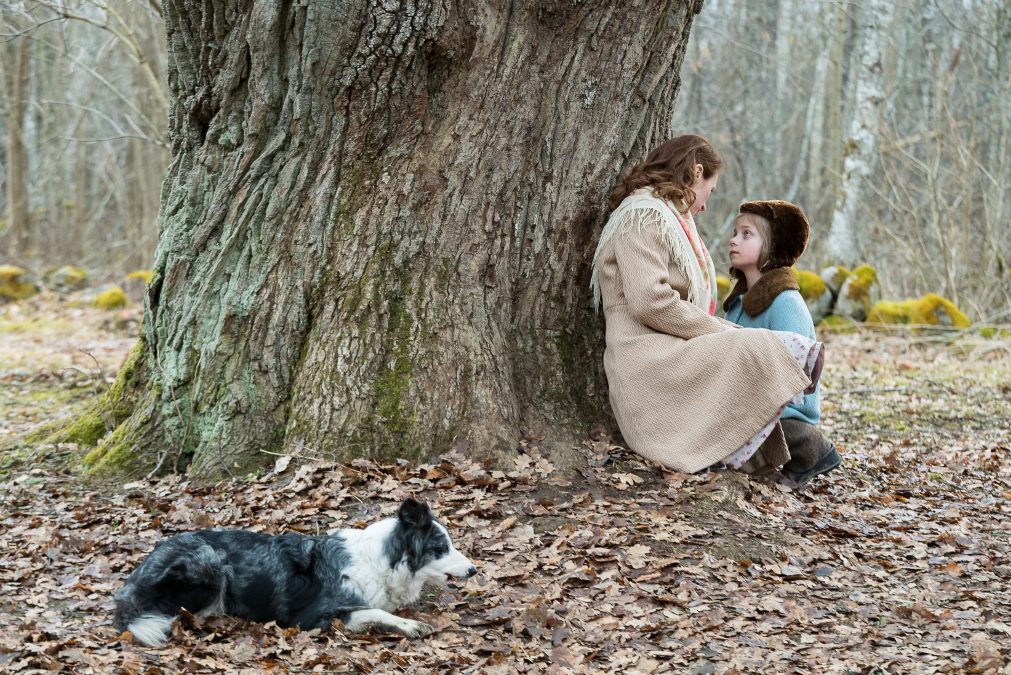 Ein Mädchen sitzt vor ihrer Mutter am Fuße eines dicken Baumes, ein Hund liegt im Vordergrund des Bildes.
