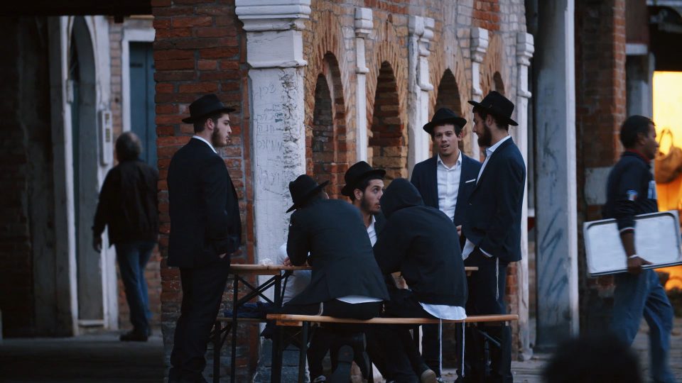 Szene aus der Dokumentation. Junge Männer mit traditionellen jüdischen Hüten stehen vor einem alten Gebäude und unterhalten sich.